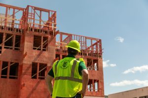 Hoover Treated Wood Products representative wearing a safety vest and hard hat standing in front of a large multi-story wood-framed building under construction demonstrating sustainable building materials in action.