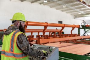 A Hoover Treated Wood Products employee in a high-visibility vest and hard hat operates a control panel at the lumber stacker on the production floor.